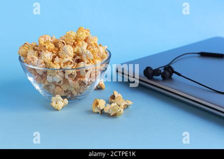 Una ciotola trasparente di popcorn su una superficie blu. Accanto a un computer portatile e alle cuffie. La famiglia che guarda i film. Spazio di copia. Foto Stock