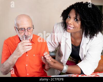 Ritratto orizzontale di un'infermiera afroamericana che tiene le mani con un uomo edero e facendolo bere un bicchiere di acqua Foto Stock