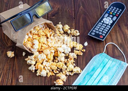 Una ciotola trasparente di popcorn su una superficie di legno. Accanto a un computer portatile e alle cuffie. La maschera protegge dai virus. Spazio di copia. Foto Stock