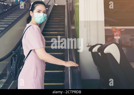 Asian Woman Hand tiene il corrimano della scala mobile, tosse nel suo gomito e mantenere la distanza sociale da altre persone con la protezione chirurgica maschera viso - lei c Foto Stock