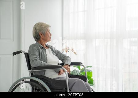 Vista posteriore di una donna anziana seduta su una sedia a rotelle che guarda fuori dal finestrino. Donna anziana nella stanza dell'ospedale seduta vicino alla finestra e pensando. Pazienti anziani Foto Stock