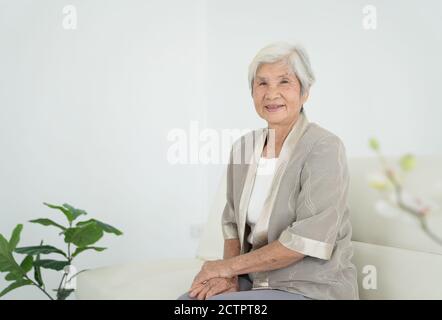 Donna anziana sorridente seduta sul divano e guardando la macchina fotografica. Svegliate la vecchia donna con i capelli grigi e i pigiami alla luce del mattino presto. Ritratto di sambuco Foto Stock