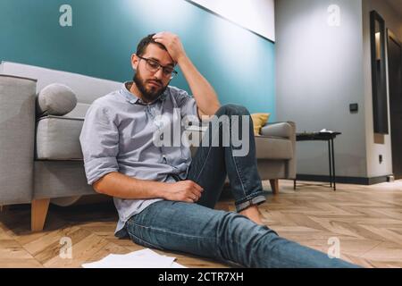 Giovane uomo confuso analizzando le finanze a casa, tenendo la testa con la mano Foto Stock