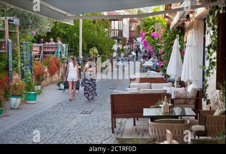 Due giovani Signore (modello rilasciato) passavano oltre il bar del Giardino Botanico lungo una stretta strada acciottolata nella Città Vecchia di Kalkan in Turchia. Kalkan è un Foto Stock