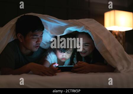 Asian Happy famiglia madre, padre e piccola figlia guardando film o cartoni animati in smartphone insieme e coperta coprire la testa a letto di notte a. Foto Stock