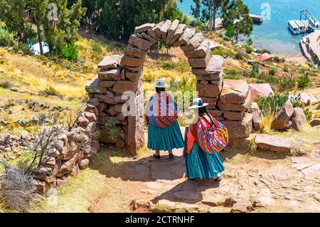 Due donne peruviane indigene Quechua in abbigliamento tradizionale sotto un arco che scende a piedi verso un porto, l'isola Taquile, lago Titicaca, Perù. Foto Stock