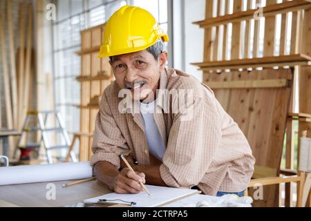 Felice anziano asiatico con casco di sicurezza che lavora in officina di Falegnameria. Concetto di persone anziane che fanno hobby dopo il pensionamento. Foto Stock
