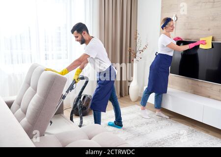 Pulitori professionali in uniforme blu piano di lavaggio e asciugatura la polvere dall'arredamento del soggiorno dell'appartamento. Il servizio di pulizia concept Foto Stock