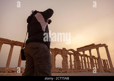 Un giovane fotografo che indossa un impermeabile e pantaloni di cotone scatta una foto delle rovine storiche dell'antica città di Palmyra all'alba. Montante in pietra Foto Stock