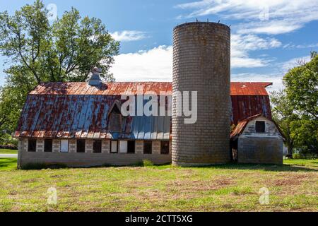 Un'antica casa colonica abbandonata con un alto silo di mattoni e un grande fienile con soffitto di stagno arrugginito. Questo antico complesso di edifici in discesa e' circondato da gr Foto Stock