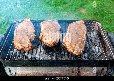 Primo piano immagine isolata di tre pezzi di bistecca di ferro piatto marinato su una griglia a carbone. La piccola griglia da picnic pieghevole è molto calda e viene fumo Foto Stock