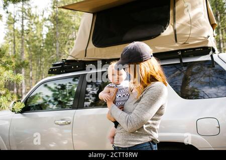 Donna che tiene figlio del bambino (6-11 mesi) sul campeggio, auto con tenda in background, Wasatch cache National Forest Foto Stock