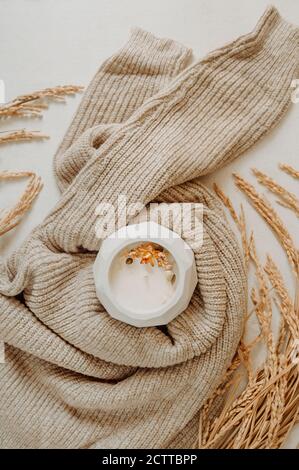 candela bianca in vaso di cemento avvolto in un accogliente pullover in maglia beige su sfondo bianco con fiori secchi, fuoco selettivo Foto Stock