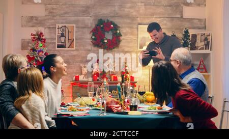 Uomo allegro che scatta una foto della sua bella famiglia alla riunione di natale. Tradizionale cena di natale festiva in famiglia multigenerazionale. Gustando la festa del pasto di Natale in una sala decorata. Grande riunione di famiglia Foto Stock