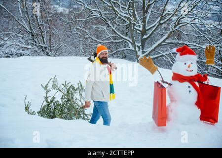 Il giovane lumberjack porta l'abete sullo sfondo bianco della neve. Un bel legname con una barba porta un albero di Natale. Giovane uomo lumberjack sta tagliando Foto Stock