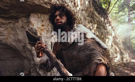 Caveman primordiale che indossa la pelle animale tiene il martello di pietra guarda intorno alla foresta preistorica, pronto a caccia di Prey animale. Neanderthal andare Caccia in Foto Stock