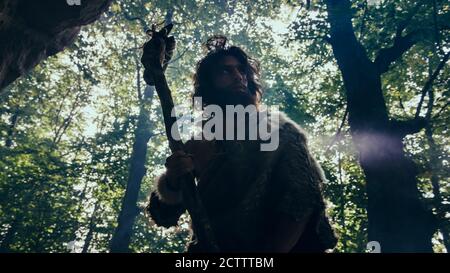 Silhouette di Primeval Caveman che indossa pelle animale tiene pietra Hammer guarda intorno Prehistoric Forest, Ready to Hunt Animal Prey. Neanderthal andare Foto Stock