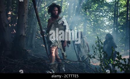 Tribù di cacciatori-raccoglitori indossando animale Skin Holding Stone Tipped Tools, esplorare Preistic Forest in a Hunt for Animal Prey. Famiglia Neanderthal Foto Stock