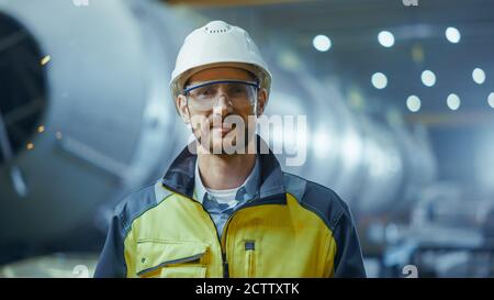 Ritratto di sorridente lavoratore professionista dell'industria pesante che indossa divisa di sicurezza, occhiali e casco. In background non messo a fuoco grande Foto Stock