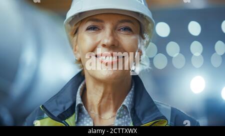 Ritratto di professionista Heavy Industry Female Engineer indossando divisa di sicurezza e Hard Hat, sorridente affascinante. In background non messo a fuoco grande Foto Stock