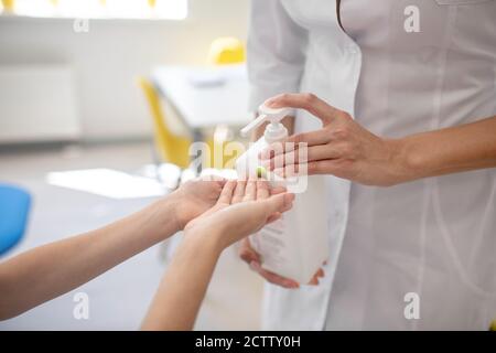 Medico in un camice da laboratorio spruzzando disinfettante alle mani dei bambini Foto Stock