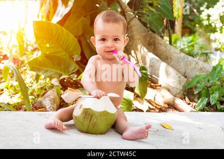 Felice bambino neonato in estate vacanza tropicale. Mangia e beve di cocco verde giovane. Siede su un terreno e tiene beva paglia e cucchiaio. Giorno di sole Foto Stock