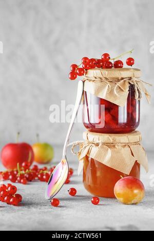 due vasetti di marmellata di frutta fatta in casa. Vasetti di vetro con diversi tipi di marmellata e bacche. Confettura di prugne e ribes rosso su sfondo grigio chiaro. Foto Stock