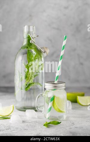 Bottiglia di vetro e vaso di limonata con limonata fredda con foglie di menta fresca e lime con cubetti di ghiaccio su fondo grigio di cemento. Fotografia alimentare Foto Stock