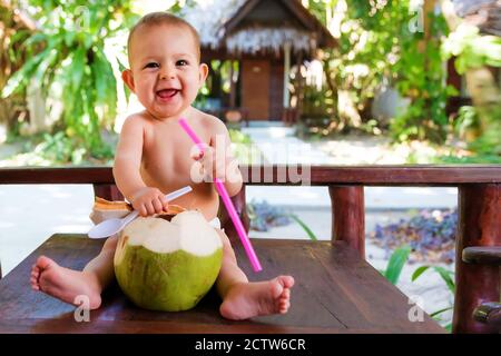 Bambino felice del bambino in vacanza tropicale. Mangia e beve di cocco verde giovane. Siede su un tavolo di legno Foto Stock