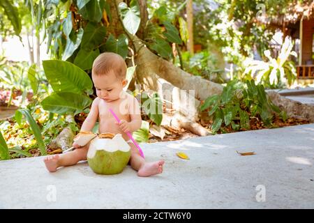 Il bambino pensivo e appassionato si siede sulla terra e. mangia e beve di cocco verde giovane con cucchiaio Foto Stock