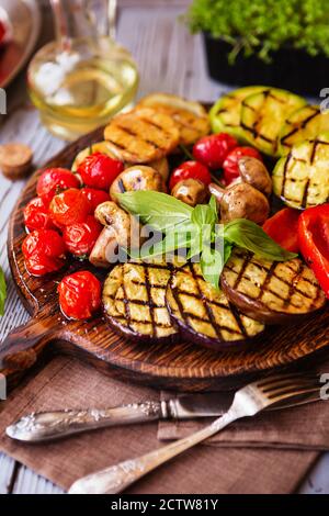 Verdure grigliate sul tagliere su sfondo di legno. Verdure alla griglia (peperone colorato, pomodori, cipolla, zucchine, melanzane) con basilico e basilico Foto Stock