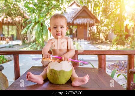 dentizione e salivazione bambino infante in vacanza tropicale. Mangia e beve di cocco verde giovane. Siede su un tavolo Foto Stock