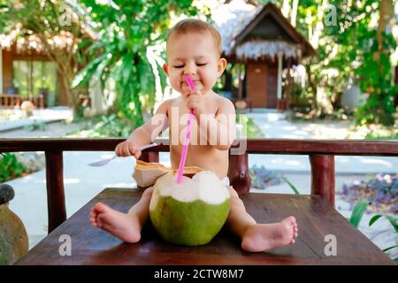 Bambino infante in vacanza tropicale. Mangia e beve di cocco verde giovane. Siede su un tavolo di legno e tiene la paglia potabile Foto Stock