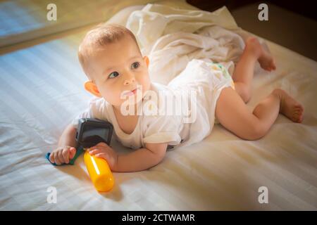 Il bambino piccolo 8 mesi giace su un letto bianco. La ragazza del bambino gioca con la macchina fotografica di azione e guardando la mamma Foto Stock