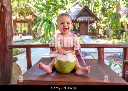 Felice bambina in vacanza tropicale. Mangia e beve di cocco verde giovane. Siede su un tavolo di legno Foto Stock