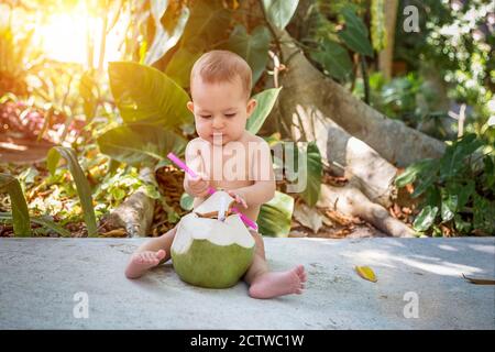 Bambino infante in vacanza tropicale. Raccoglie e beve giovane cocco verde con paglia potabile Foto Stock