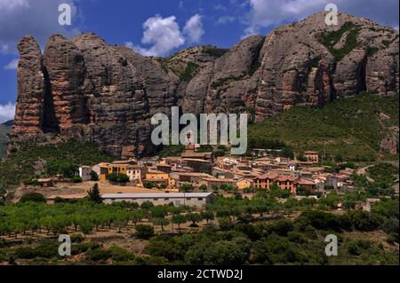 Agüero, un piccolo villaggio ai piedi dei Pirenei a Huesca, Aragona, Spagna, si aggira intorno ad una bella chiesa romanica del 13 ° secolo e si accoccola sotto Los Mallos de Agüero (i Mallets o Ninepins di Agüero). Questa spettacolare formazione, conosciuta anche come le colline di Syncline, è costituita da una roccia conglomerata di epoca terziaria erosa dalle tinte rosse che sale fino a 900 piedi / 275 metri sopra l'insediamento. Foto Stock