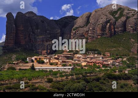 Agüero, un piccolo villaggio ai piedi dei Pirenei a Huesca, Aragona, Spagna, si aggira intorno ad una bella chiesa romanica del 13 ° secolo e si accoccola sotto Los Mallos de Agüero (i Mallets o Ninepins di Agüero). Questa spettacolare formazione, conosciuta anche come le colline di Syncline, è costituita da una roccia conglomerata di epoca terziaria erosa dalle tinte rosse che sale fino a 900 piedi / 275 metri sopra l'insediamento. Foto Stock