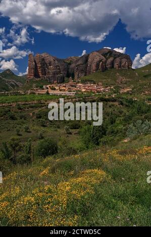 Vista a distanza di Agüero nella provincia di Huesca, Aragona, Spagna, un piccolo villaggio ai piedi dei Pirenei dwarfed dalle colline di Syncline o Los Mallos de Agüero (i Mallets o Ninepins di Agüero). Questa spettacolare formazione di roccia conglomerata di epoca terziaria dalle tinte rosse, scolpita dall'erosione del vento e dell'acqua, sorge fino a 275 metri (900 piedi) sopra l'insediamento. Il villaggio si raggruppa intorno ad una bella chiesa romanica 13 ° secolo. Foto Stock