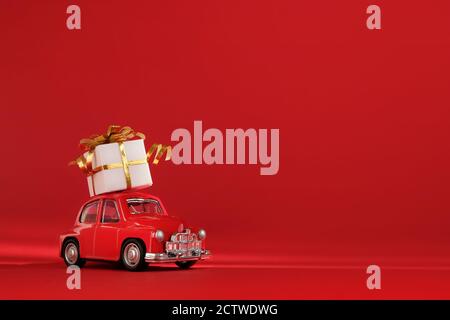 Piccola auto giocattolo retrò rossa con regalo sul tetto su sfondo rosso. Consegna. Capodanno, Natale, San Valentino, Giornata Mondiale delle Donne, Vendita. Foto Stock