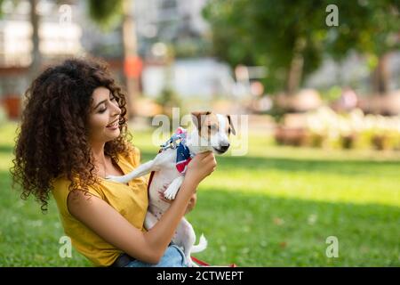 Fuoco selettivo della giovane donna sull'erba con il cane dentro bandana di bandiera americana Foto Stock