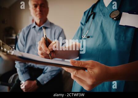 Closeup delle mani di una giovane dottoressa che scrive medico note sulla clipboard con paziente senior in background Foto Stock