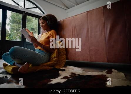 Giovane donna soddisfatta in abiti casual e cuffie wireless surf tablet digitale seduto sui cuscini del divano Foto Stock