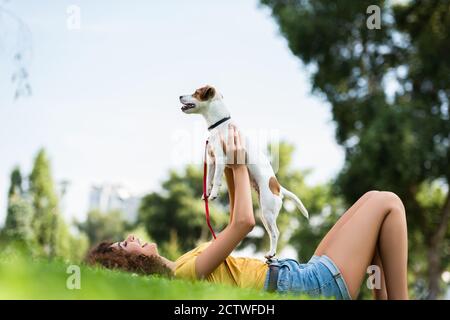 vista a livello della superficie di una donna entusiasta nella tenuta estiva dell'outfit jack russell cane terrier mentre si trova su erba nel parco Foto Stock