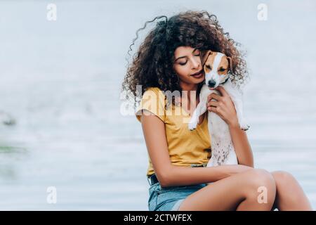 curly donna in estate vestito hugging jack russell terrier cane vicino al lago Foto Stock