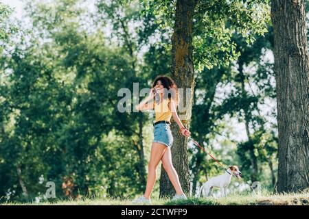 donna entusiasta in outfit estivo che parla sullo smartphone mentre cammina con il cane russell terrier jack sul guinzaglio Foto Stock