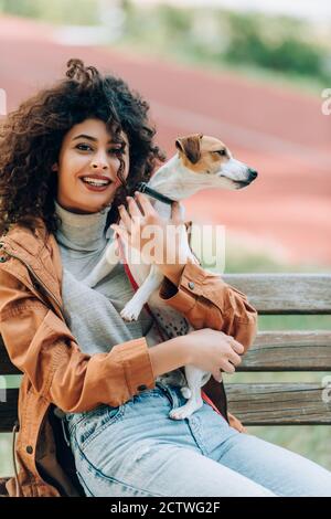 donna curly in abito autunnale guardando la macchina fotografica mentre abbracciando jack russell cane terrier su panca in parco Foto Stock