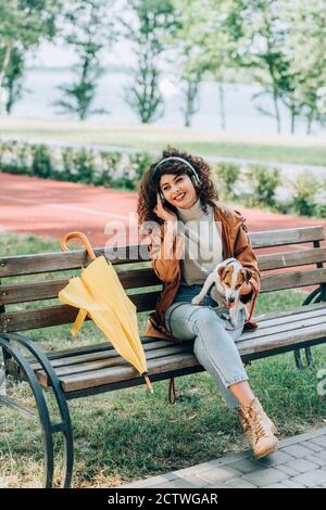 donna elegante con cuffie wireless che tengono jack russell terrier cane mentre si siede sulla panchina vicino all'ombrello giallo Foto Stock