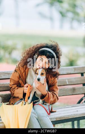 curly donna in cuffie wireless hugging jack russell terrier cane mentre si siede sulla panchina con ombrello giallo Foto Stock