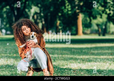 Curly donna in impermeabile hugging jack russell terrier sul prato nel parco Foto Stock